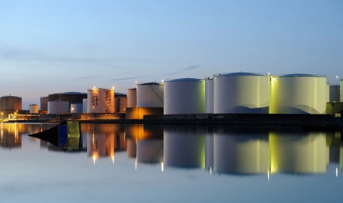 Large cylindrical industrial tanks at a harbour at dusk, lit up and reflected on the calm surface of the water.