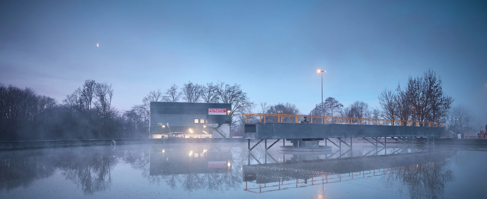 An industrial plant partly hidden by mist next to a water basin at dawn, a lit-up building with the FILTRATEC logo, a jetty construction with reflections in the calm water, moon in the sky