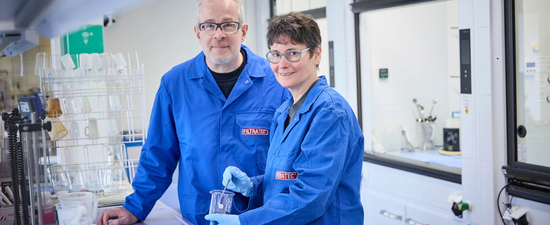 Two employees in blue FILTRATEC lab coats working together in the laboratory holding a beaker containing a sample.