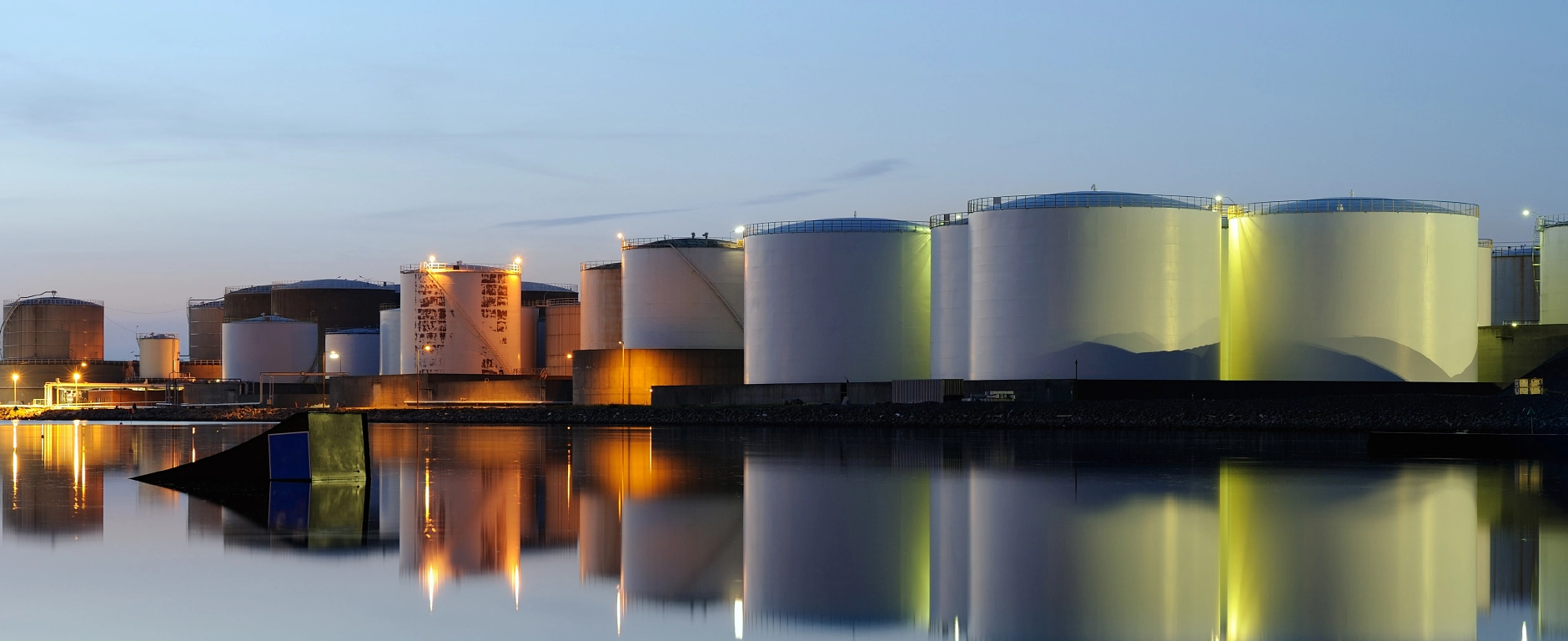 Large cylindrical industrial tanks at a harbour at dusk, lit up and reflected on the calm surface of the water.
