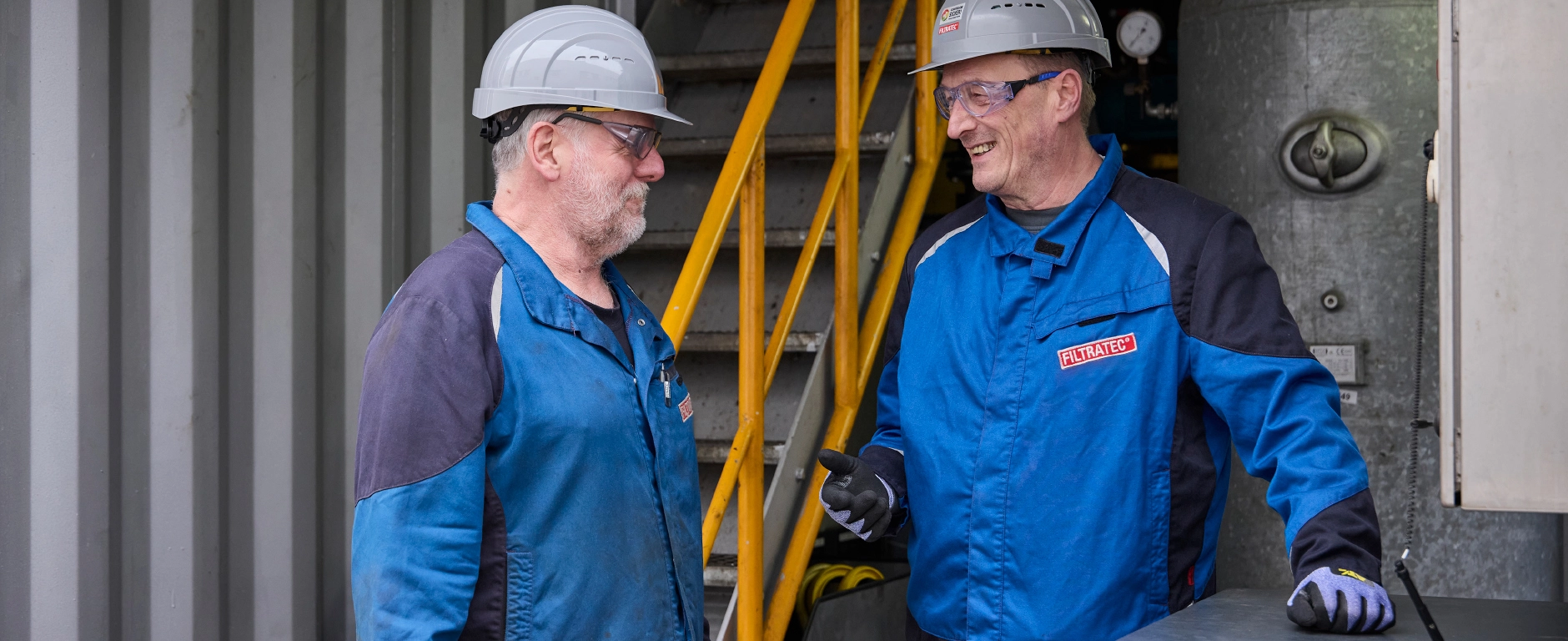 Two employees wearing FILTRATEC protective clothing and hard hats talking to each other in a decanter.