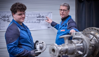 A master engineer using a technical drawing to explain a metal part to an apprentice in a workshop.