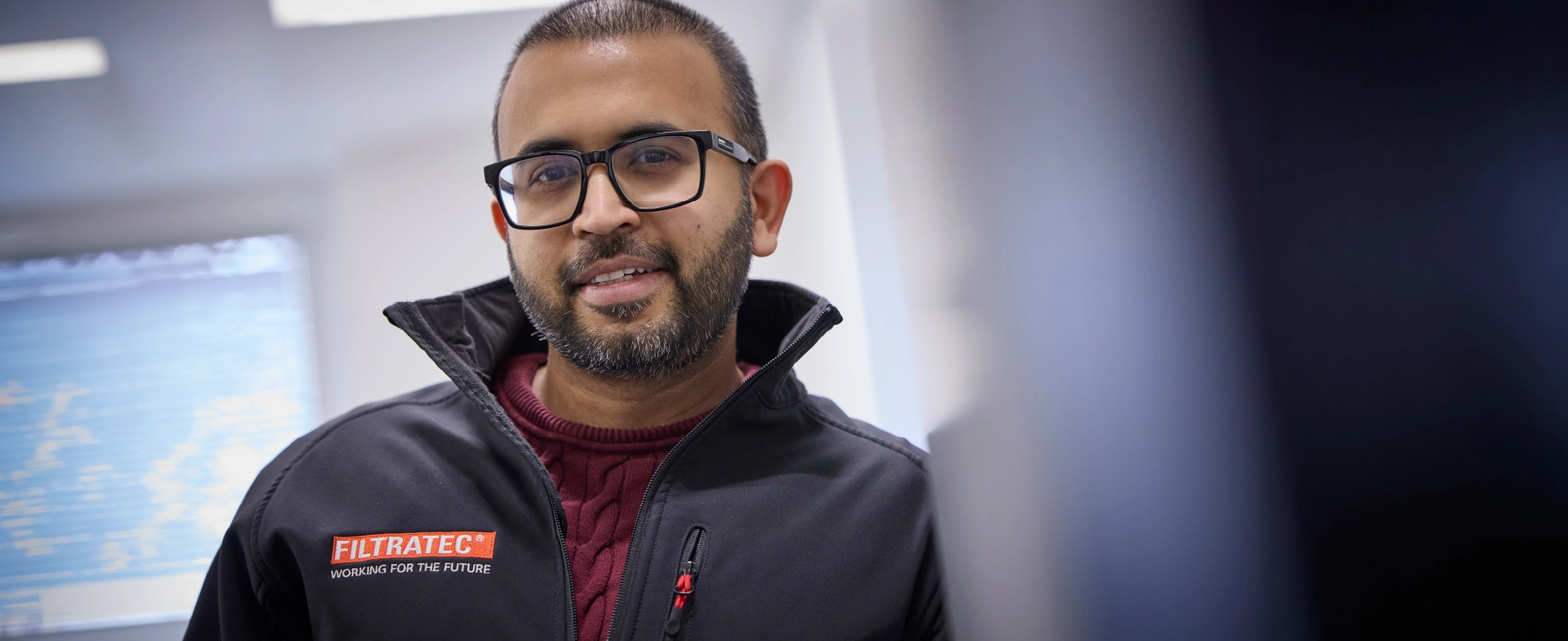 An employee wearing glasses and a FILTRATEC jacket and smiling, a portrait focusing on his face and the company logo.