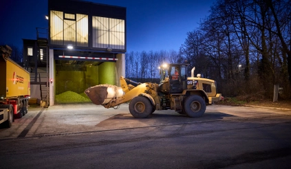 A wheel loader loading filter cake onto a waiting lorry in front of a stationary filter press on a business premises at dusk.