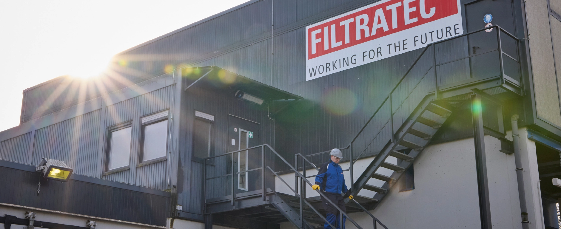 An industrial building with a FILTRATEC sign and ‘Working for the Future’ slogan, an employee wearing protective clothing on an outside staircase in the sunlight.